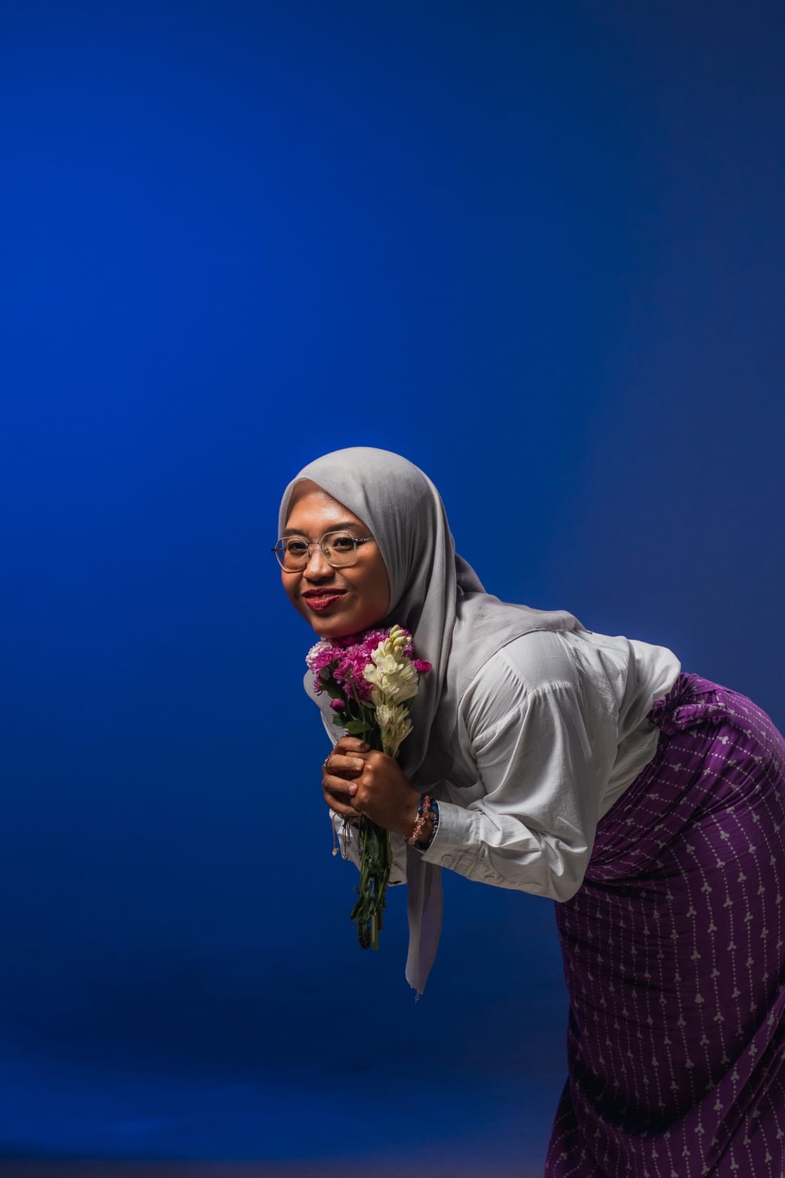 Gita Swasti leaning forward with a bouquet of pink and white flowers against a blue backdrop